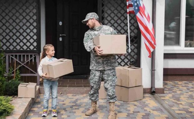 father in military uniform and daughter holding cardboard boxes near house with American flag