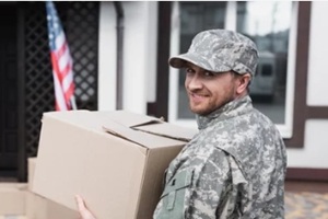 happy military serviceman holding cardboard box and looking at camera with blurred girl on background