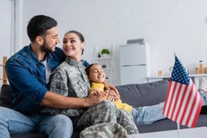 positive man hugging wife in camouflage uniform near daughter and blurred american flag at home